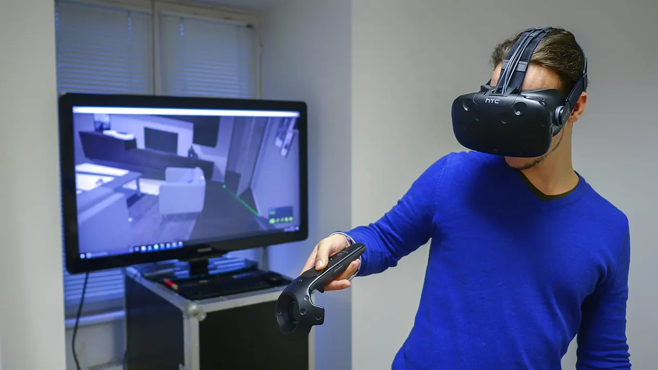 A man with an HTC Vive headset in front of an apartment walk-through app, the monitor showing the 3D view of the flat